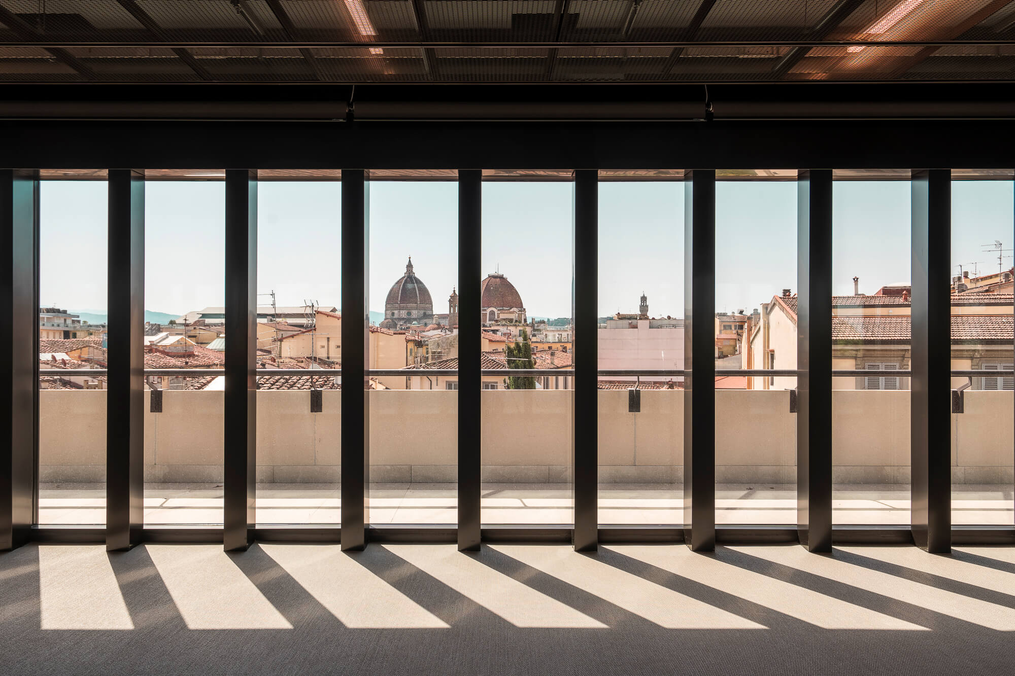 Vista attraverso grandi vetrate di una città, con i tetti e le cupole di edifici storici, tra cui una grande cupola visibile in background.