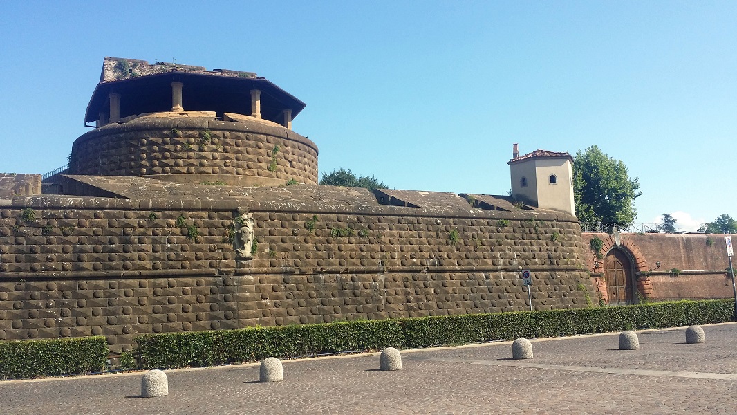 Vista di una fortezza in muratura con una cupola, caratterizzata da un muro decorato con motivi circolari e una faccia scolpita. Si vedono anche delle piante che crescono sulla superficie. Sullo sfondo, un edificio con un tetto rosso e alberi verdi.