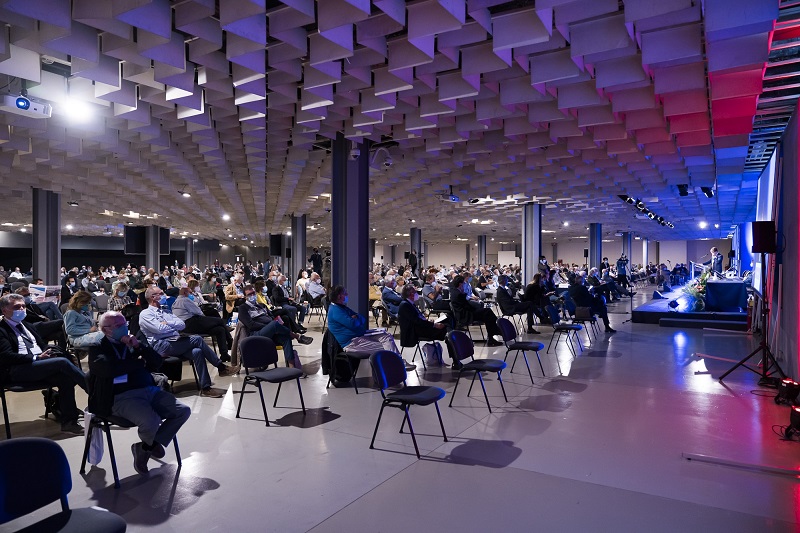 Un'ampia sala conferenze con un pubblico numeroso di partecipanti seduti su sedie, alcuni indossano mascherine. Sul palco due oratori presentano, con luci colorate che illuminano l'area; in fondo ci sono schermi e attrezzature audio.