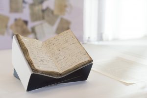 Un libro aperto con pagine ingiallite e scritte a mano, esposto su un supporto bianco e nero, con un sfondo che mostra documenti vecchi in chiaroscuro.