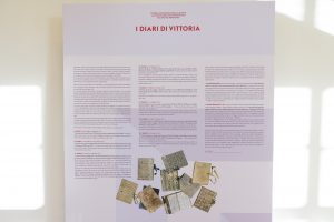 Un pannello informativo intitolato "I diari di Vittoria", con testo in italiano e una serie di diari posizionati in basso, ognuno con copertine decorate in vari stili e colori. Il pannello descrive i contenuti dei diari e il loro significato, con una grafica semplice e un layout moderno.
