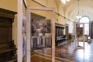 Una vista di un'esposizione in un edificio storico, con pannelli che mostrano fotografie in bianco e nero di interni, tra cui una cucina e un salotto. A sinistra, si intravede un mobile in legno elaborato e il pavimento presenta un motivo geometrico. La luce proveniente da una grande finestra illumina l'ambiente.