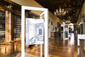 Interno di un museo con una lunga sala luminosa, arredi in legno, e opere espositive; in primo piano, un pannello bianco con un'immagine in bianco e nero di un interno storico.