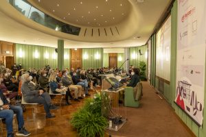 Una sala conferenze affollata con persone sedute in riga, alcune indossano mascherine. Sul palco ci sono relatori, con un monitor che mostra un'immagine e un pannello informativo a destra. Le pareti sono verdi e l'illuminazione è soffusa.