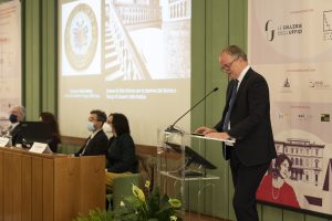 Un relatore al microfono durante una conferenza, con vari relatori seduti a un tavolo di fronte. Sullo sfondo ci sono proiezioni di immagini artistiche. Il relatore indossa una giacca nera e una cravatta blu ed è intento a parlare, mentre gli altri partecipanti, alcuni con mascherine, osservano.