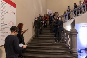 Un gruppo di persone su una scala di un museo, alcune in piedi e altre sedute, mentre una donna con capelli ricci legge un libro. Sullo sfondo è visibile una grande mappa con informazioni sui piani del museo, e ci sono cartelli informativi sulle pareti.