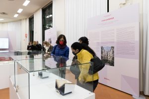 Visitatrici in un museo osservano oggetti esposti in teche di vetro, con pannelli informativi sullo sfondo che descrivono la "Casa Gardner". Una donna indossa una giacca blu, un'altra una giacca gialla, mentre altre persone si trovano in fondo alla sala.