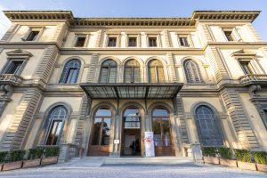 Facciata di un elegante edificio storico con finestre ad arco e una grande entrata centrale, caratterizzato da decorazioni architettoniche in pietra e doppi balconi. La vista è illuminata dalla luce del sole, con un cartello pubblicitario vicino all'ingresso e piante in vaso ai lati.