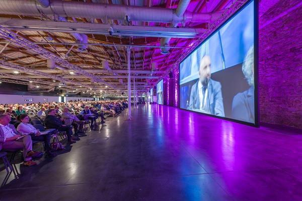Una grande sala conferenze con un pubblico numeroso seduto su sedie, illuminata da luci viola, e due schermi giganti che mostrano relatori sul palco.
