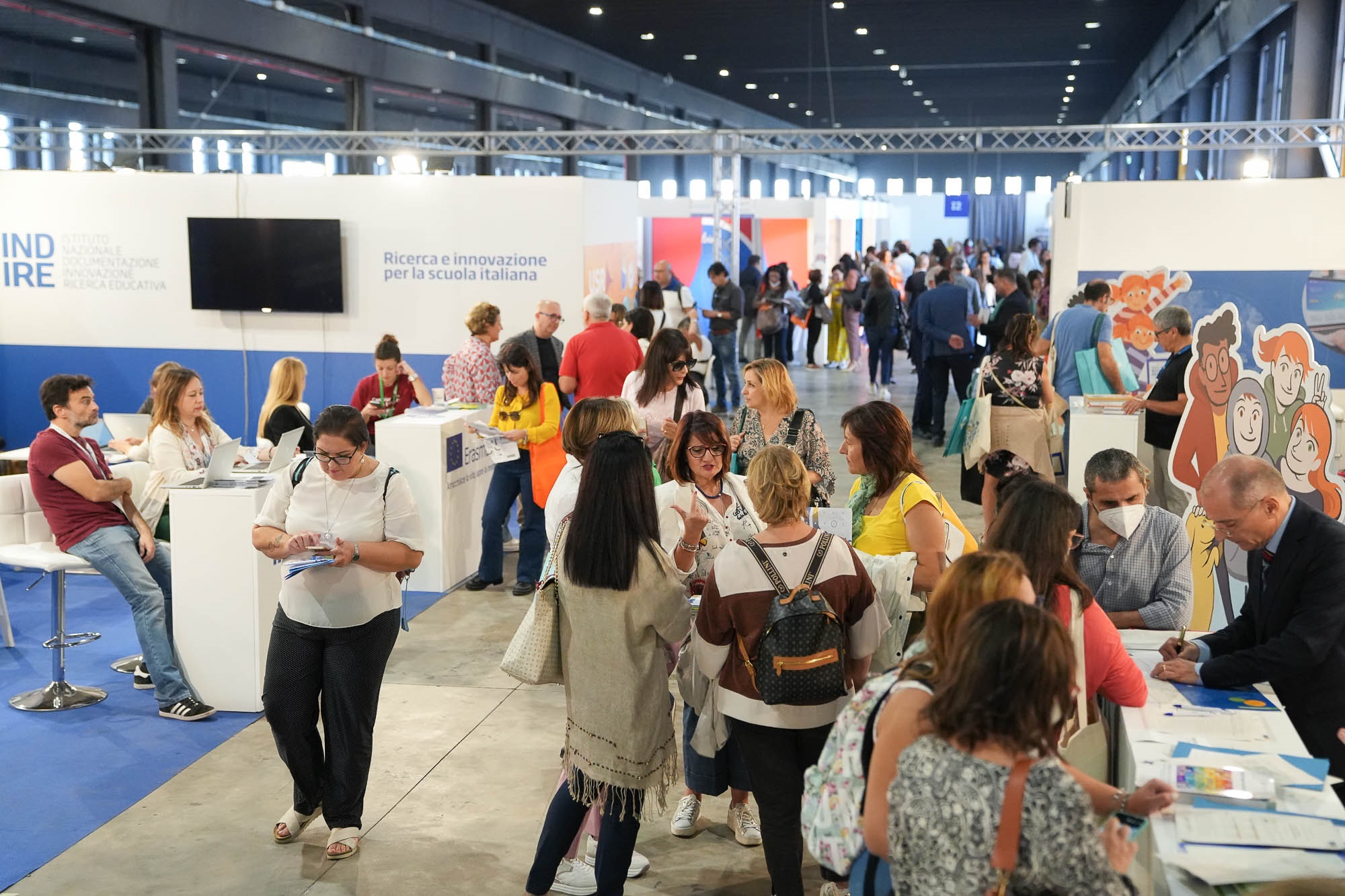 Una fiera con molte persone che interagiscono tra loro, mentre visitano vari stand educativi, inclusi alcuni con cartelloni colorati e un grande schermo. Alcuni partecipanti sono seduti e usano computer portatili, mentre altri discutono e scambiano informazioni.