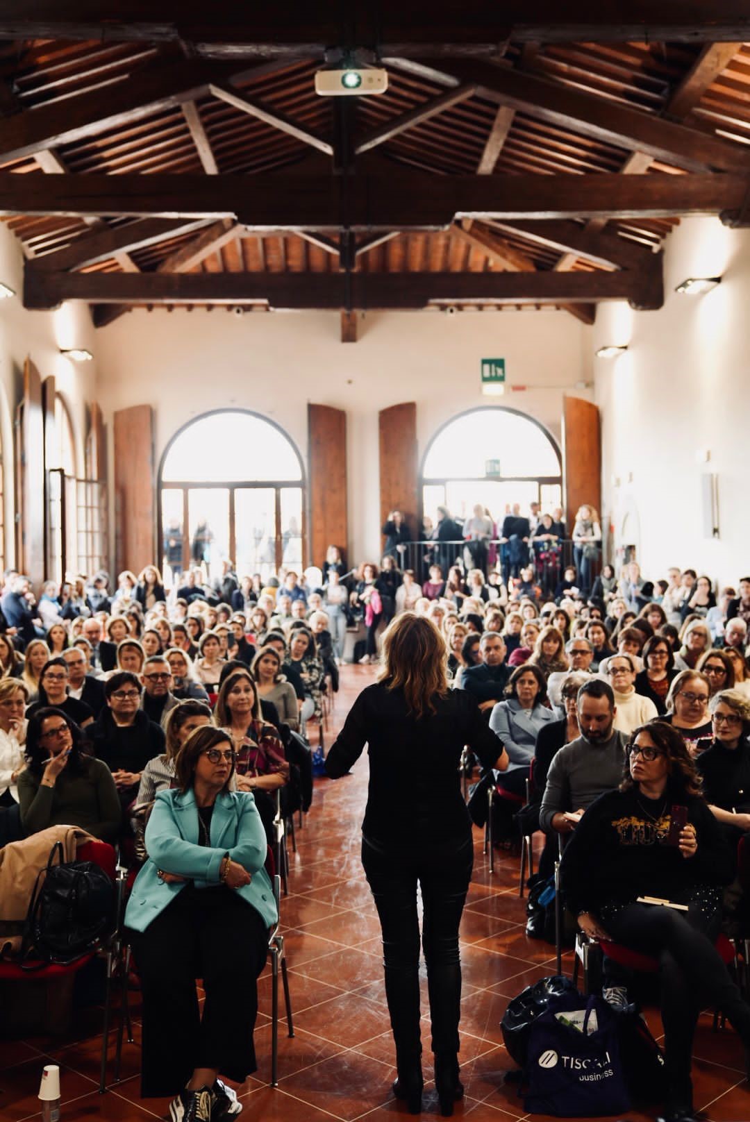 Una relatrice in piedi davanti a un ampio pubblico in una sala con soffitto a travi di legno, gli spettatori sono principalmente donne sedute, visibili in una varietà di abbigliamento, mentre alcuni si affacciano dalle finestre sul retro.