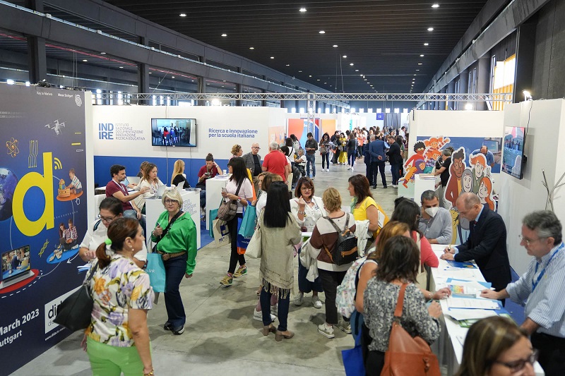 Una fiera con molte persone che interagiscono negli stand; diversi espositori presentano informazioni e materiali, con poster colorati sui muri e schermi in evidenza.