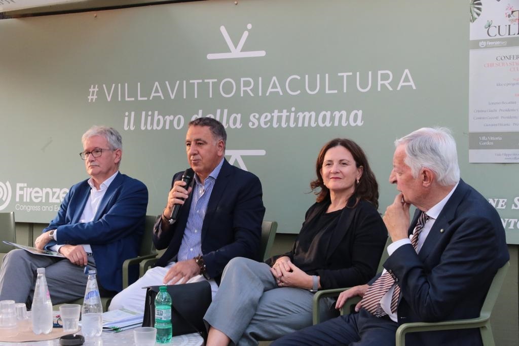 Quattro relatori seduti in un evento culturale, con uno di loro che tiene un microfono. Sullo sfondo, un grande cartello con la scritta #VILLAVITTORIACULTURA. I relatori sono in abbigliamento formale e si trovano in un ambiente all'aperto.
