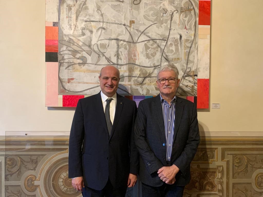 Due uomini in abiti formali sorridono insieme di fronte a un grande dipinto astratto con toni grigi, rossi e pink, all'interno di una sala con decorazioni eleganti.