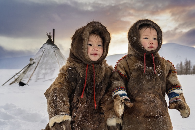 Due bambini vestiti con abiti di pelliccia, sorridenti, in piedi nella neve su uno sfondo di un paesaggio invernale, con un tepore tradizionale sullo sfondo.