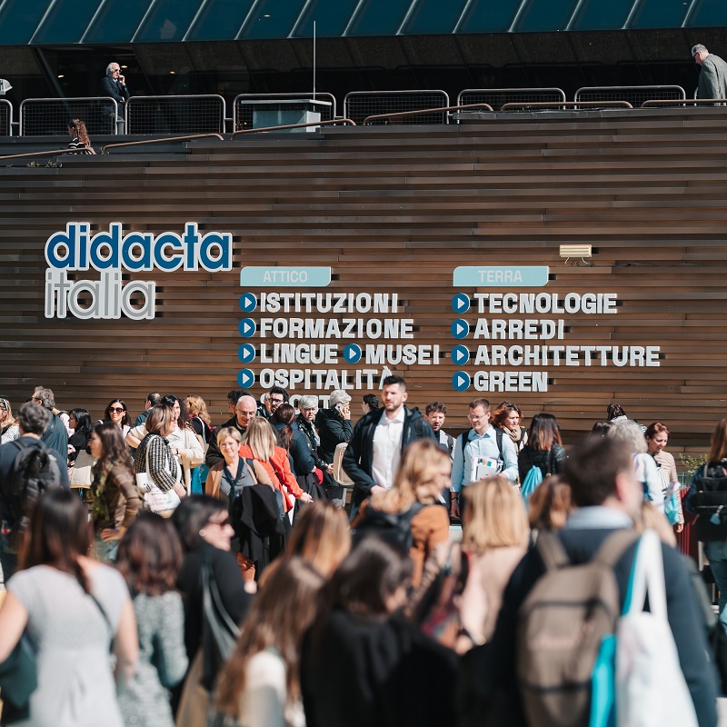Una folla di persone si raduna davanti al pannello della fiera Didacta Italia, che elenca categorie come 'Istituzioni', 'Formazione', 'Lingue', 'Musei', 'Ospitalità', 'Tecnologie', 'Arredi', 'Architetture' e 'Green'.