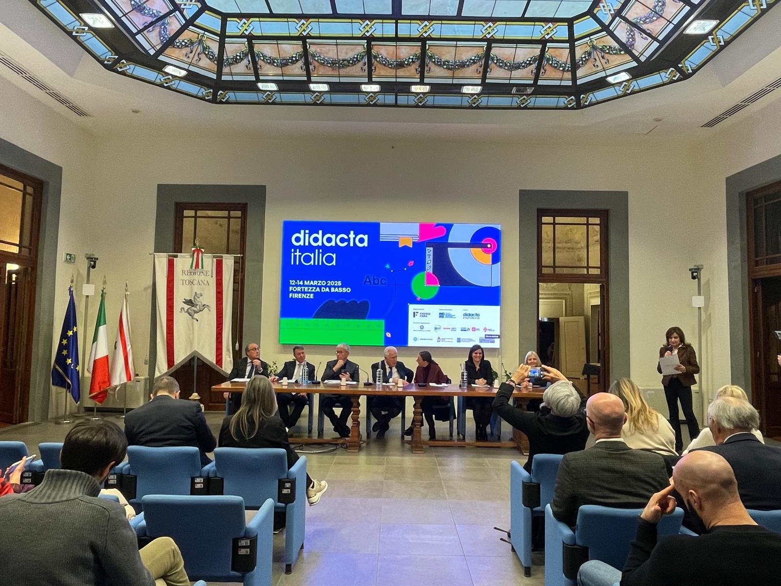 Una conferenza stampa con un tavolo di relatori e un grande schermo sullo sfondo che presenta informazioni sull'evento 'Didacta Italia', in programma dal 12 al 14 marzo 2025 a Firenze.